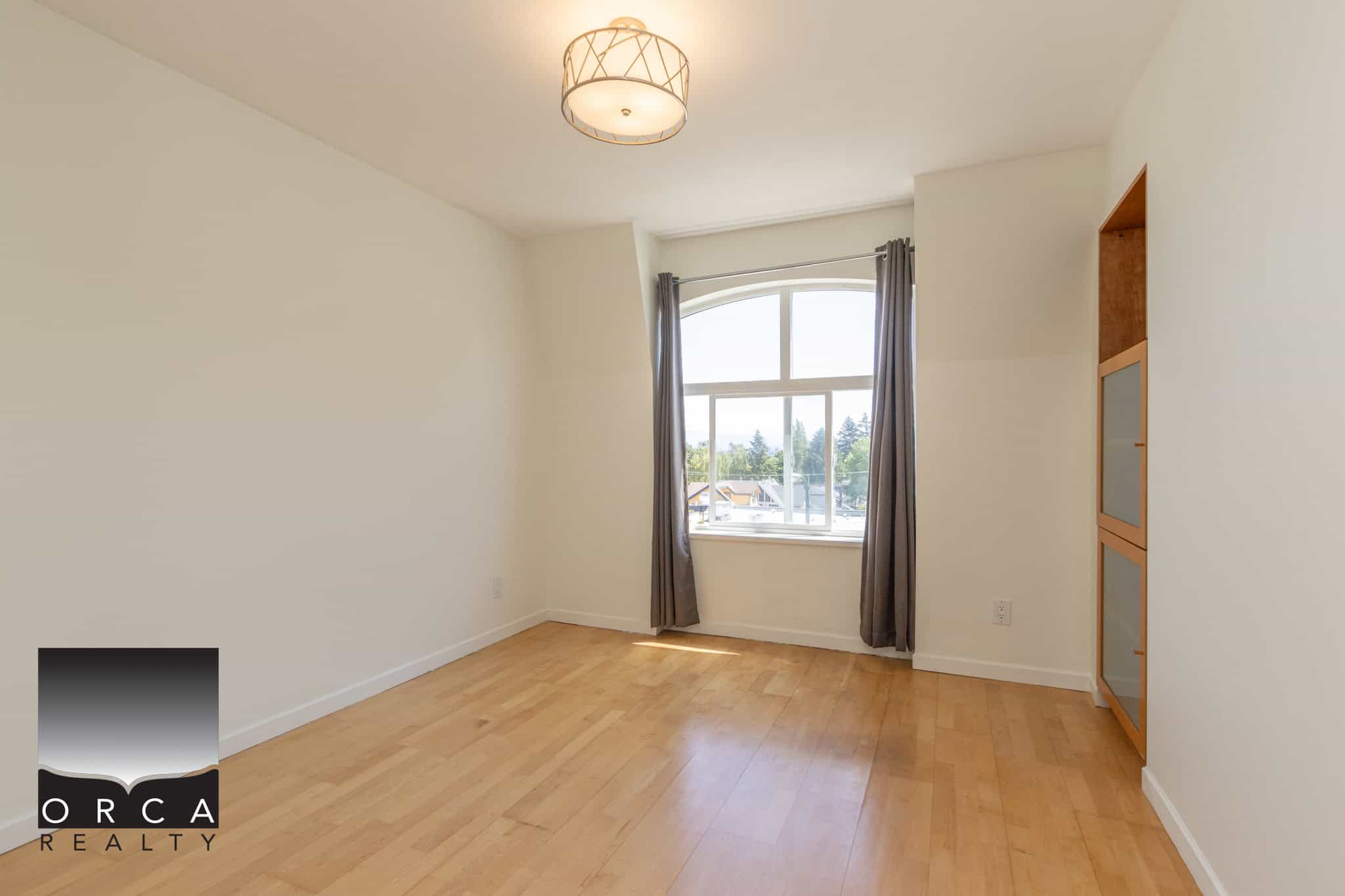 Bright empty bedroom in Vancouver condo with large arched window, natural light, hardwood flooring, modern ceiling light fixture, and built-in storage from Orca Realty Inc.