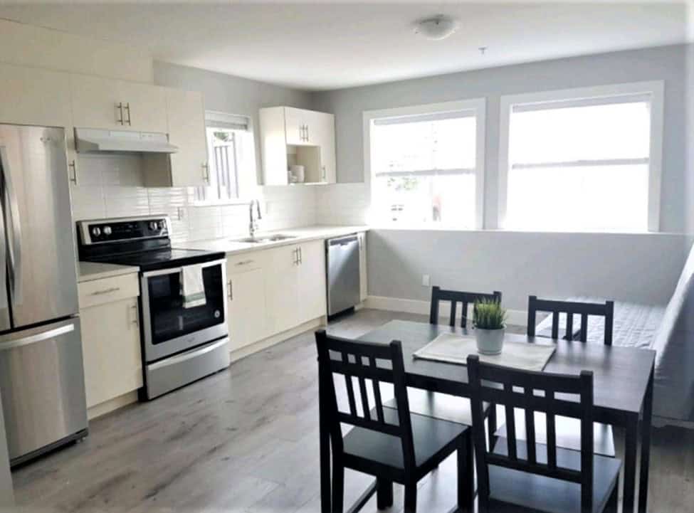 Bright modern kitchen with stainless steel appliances and white cabinetry, adjacent to dining area with black table and chairs, large windows providing natural light.