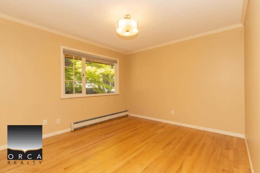 Spacious empty room with hardwood flooring, large window with lush green view, neutral beige walls, and modern ceiling light fixture. Perfect for family living or cozying up with your favorite decor.