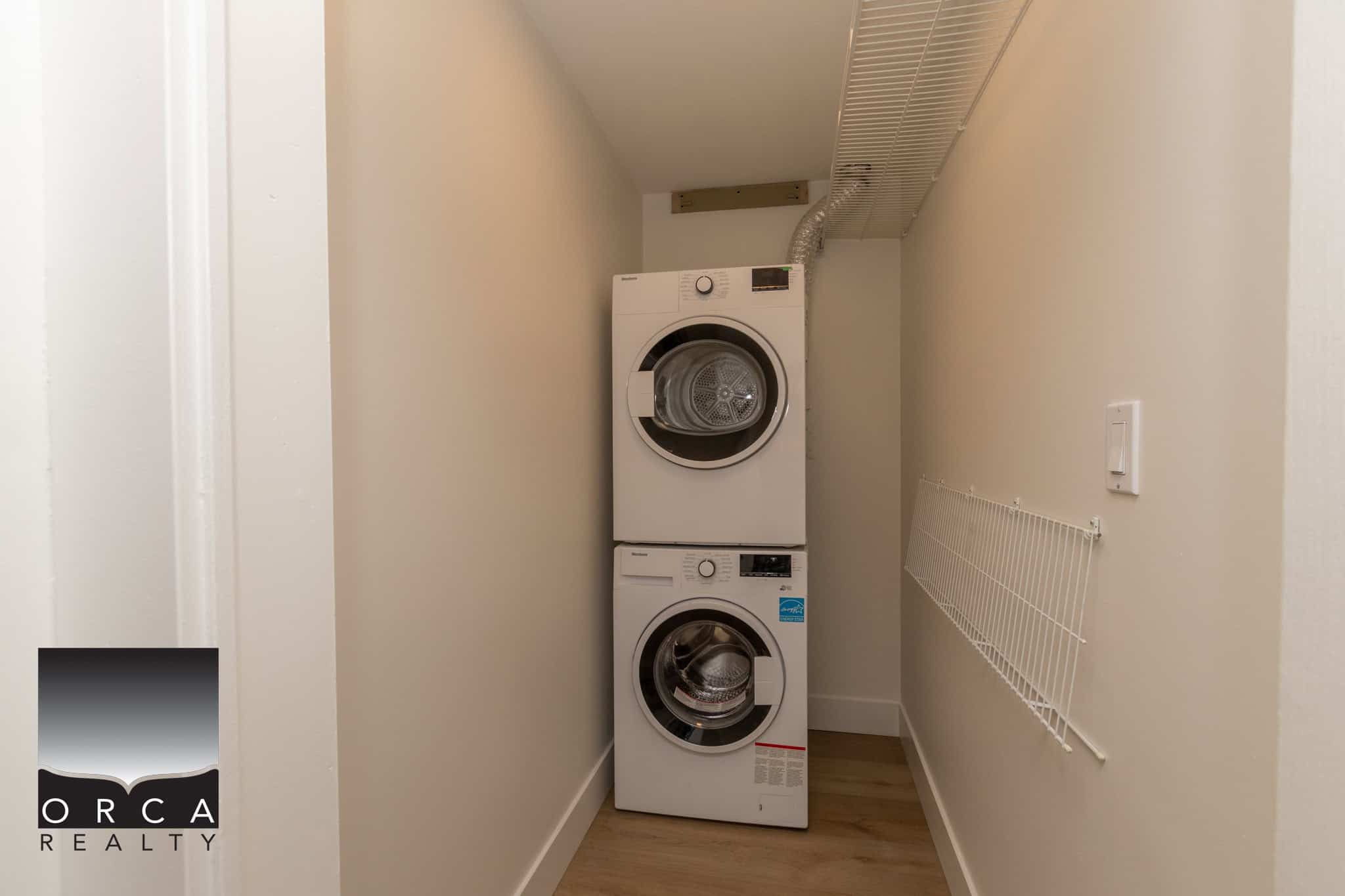 Stacked washer and dryer in a small laundry room with white walls and wood flooring, perfect for an easy laundry setup in a residential property listed by Orca Realty Inc.