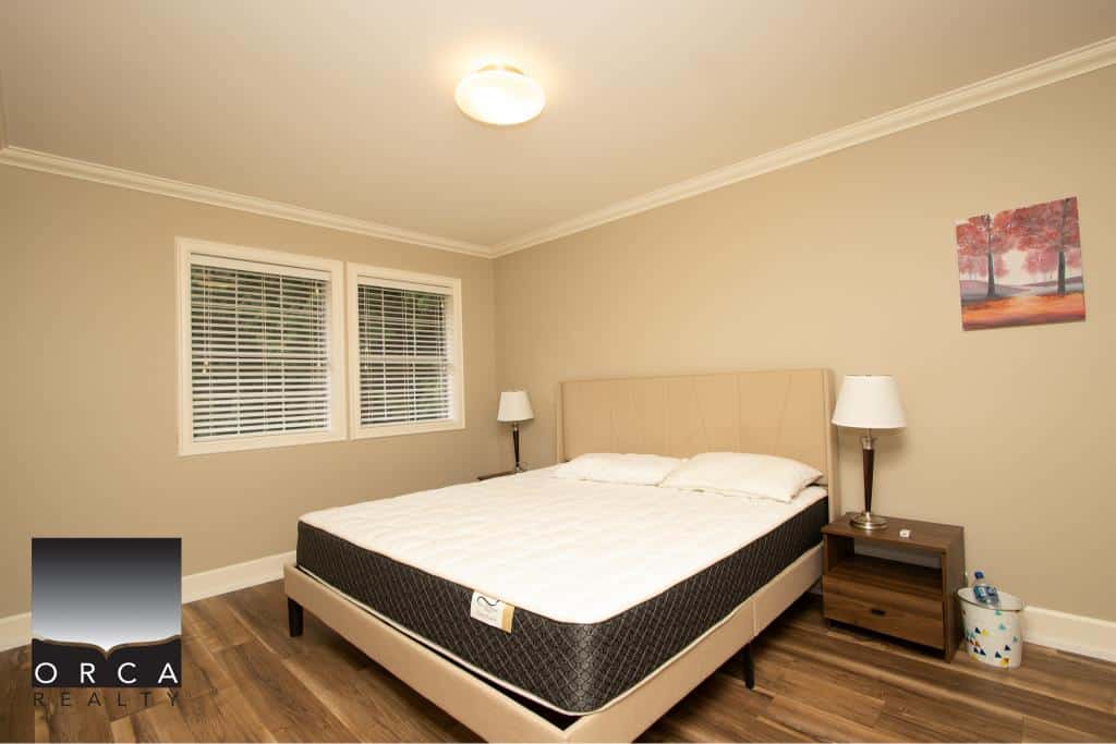 Modern bedroom with beige walls, hardwood flooring, and a large bed with white bedding, featuring two nightstands with lamps, double window blinds, and a colourful wall art piece, showcasing quality real estate listings by Orca Realty Inc.
