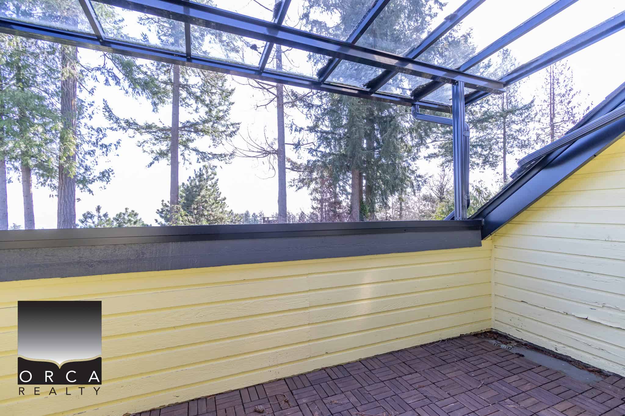 Bright yellow exterior balcony with glass roof, overlooking a forest of tall evergreen trees, offering a cozy outdoor space in a peaceful setting.