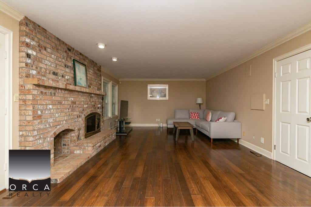 Bright spacious living room with brick fireplace, hardwood floors, neutral wall colors, and ample natural light, ideal for family gatherings and cozy evenings.