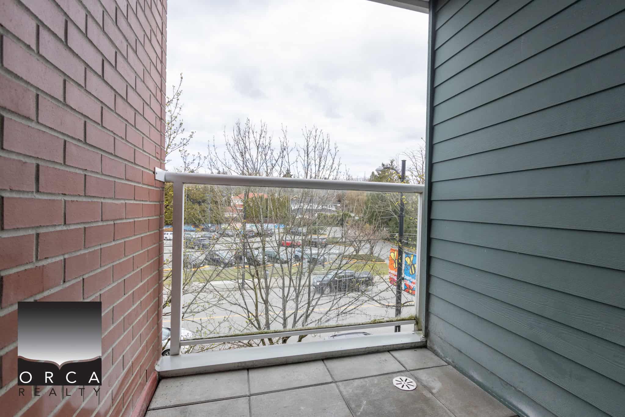 Bright balcony with glass railing overlooking parking lot and trees, ideal for relaxing or outdoor activities, part of a modern residential property listed by Orca Realty Inc. in Victoria BC.