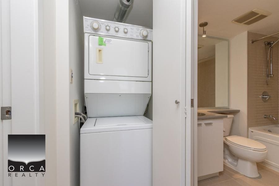 Stacked washer and dryer unit in a compact laundry area with closet doors, adjacent to a modern bathroom with a toilet, sink, and bathtub, showcasing functional home amenities.