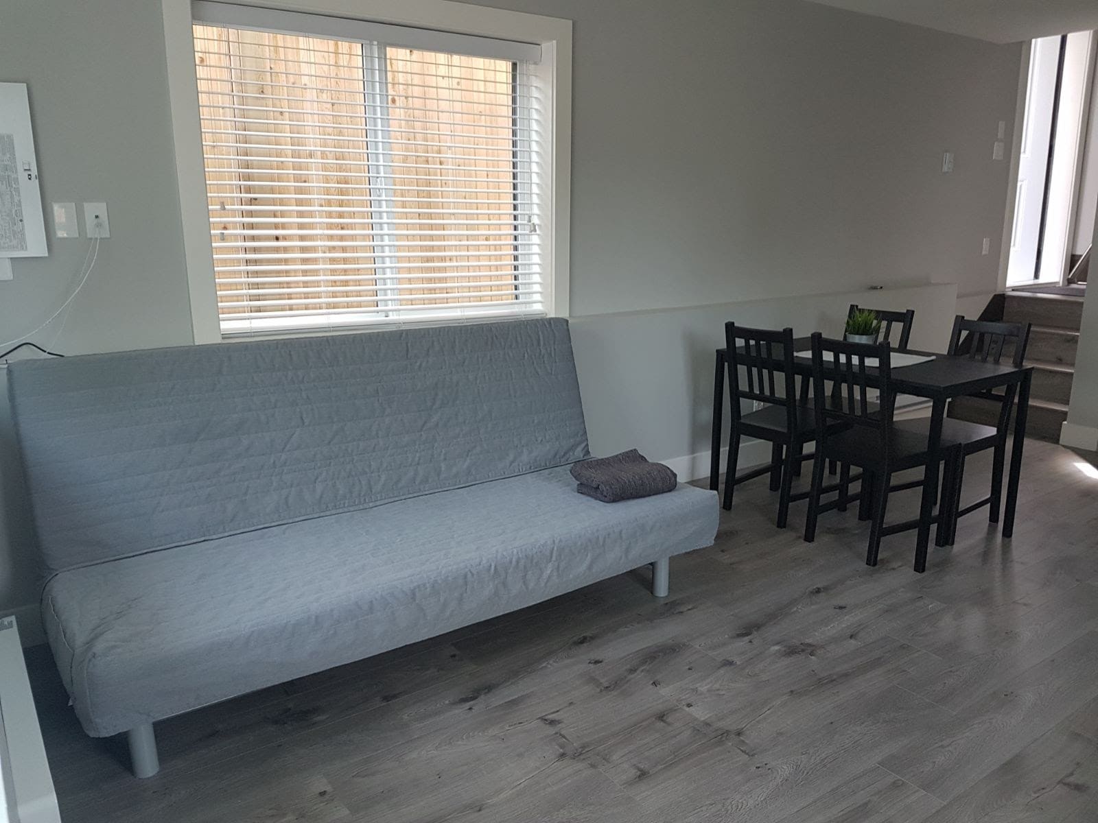 6 Modern living room with light-colored futon, window with blinds, and a black dining table set with four chairs in a bright, spacious area.