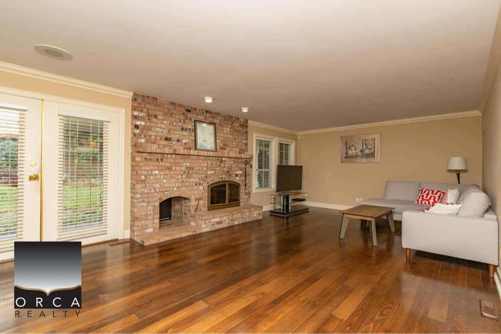 Spacious living room with brick fireplace, hardwood floors, modern TV setup, and comfortable seating area in a beautiful Orca Realty Inc. listing.