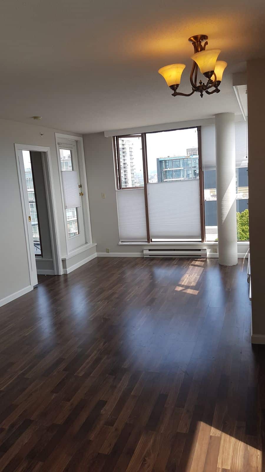 Bright modern living room with large windows offering city views, featuring hardwood floors, white walls, and a stylish ceiling light fixture, perfect for Vancouver condo living.