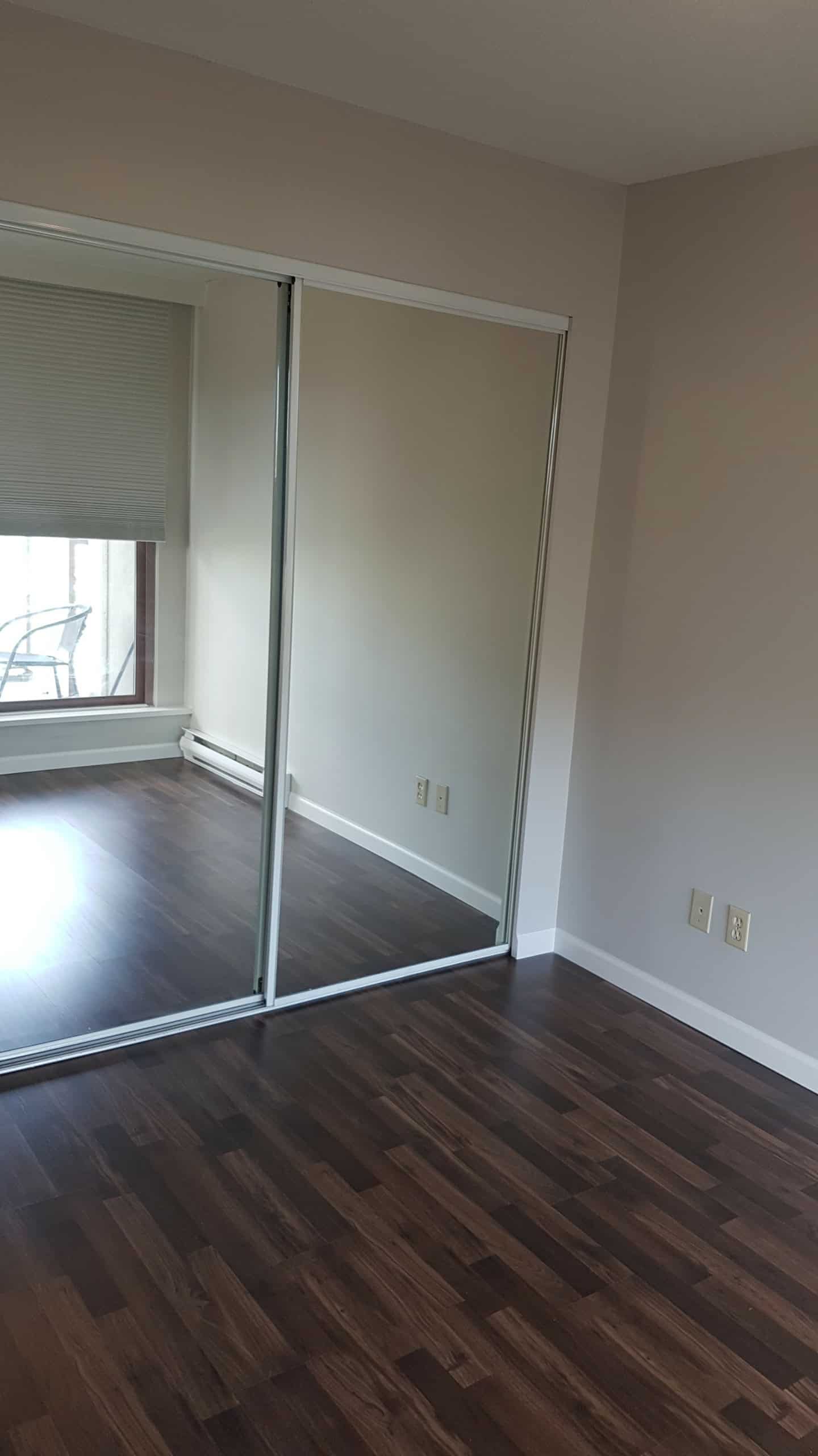 Bright empty bedroom with hardwood floors and mirrored closet doors, suitable for residential or rental purposes, featuring modern interior design, natural light from window, and clean white walls.