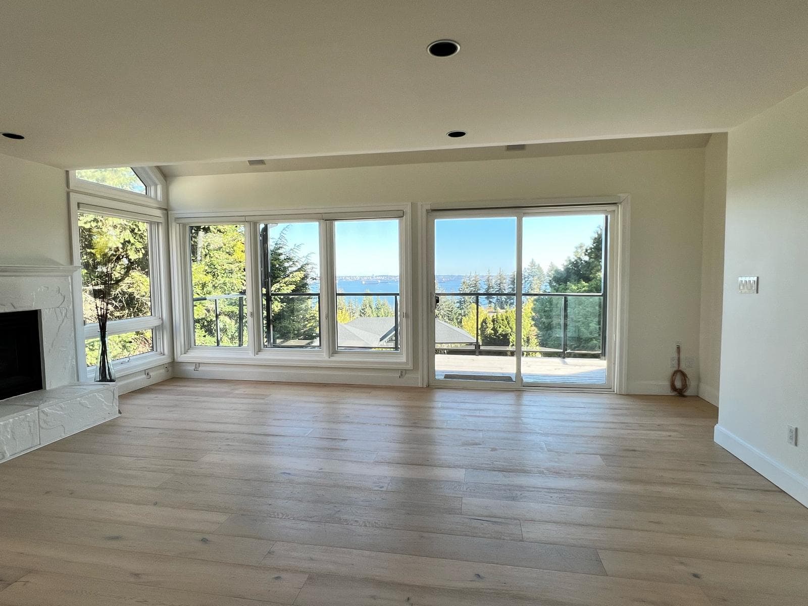 Bright living room with large windows offering panoramic ocean and forest views, hardwood flooring, and access to a balcony. Perfect space for relaxing or entertaining while enjoying nature.