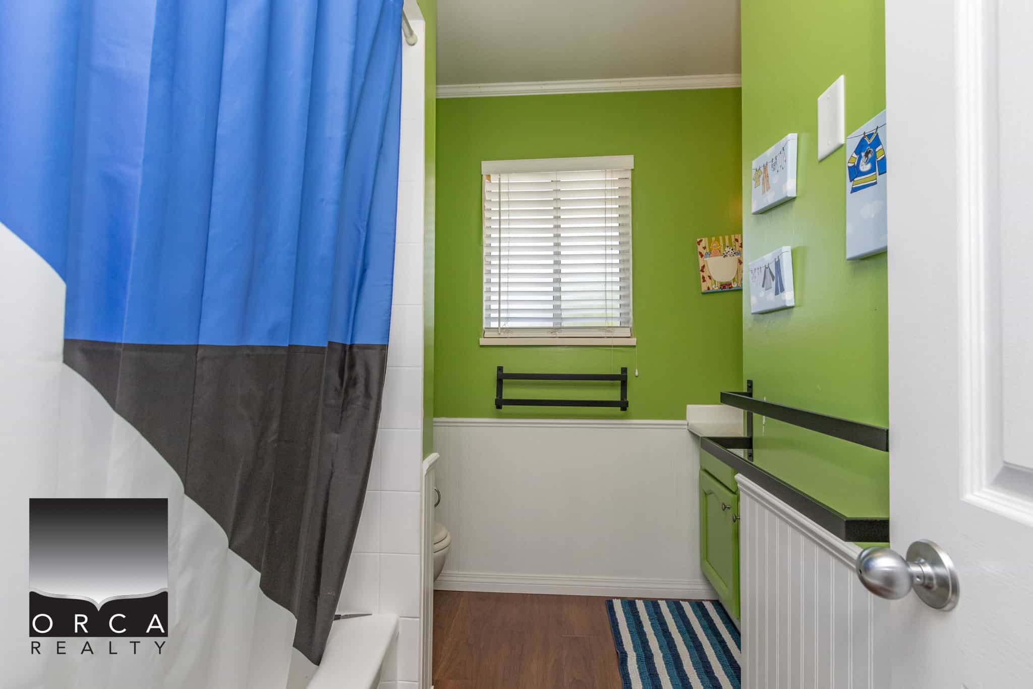 Bright green bathroom with white and black accents, featuring a striped rug, framed artwork, a small vanity, and a window with white blinds, perfect for a family home or rental property.