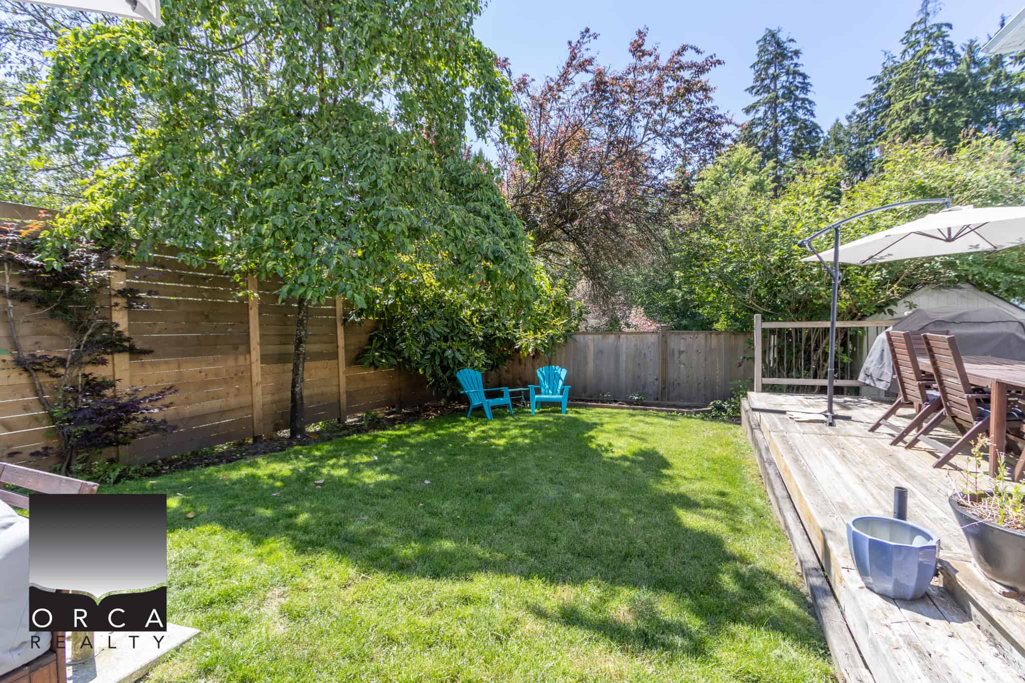Lush backyard with green grass, mature trees, and outdoor seating area, perfect for relaxing or entertaining, showcasing ideal outdoor living space offered by Orca Realty Inc.