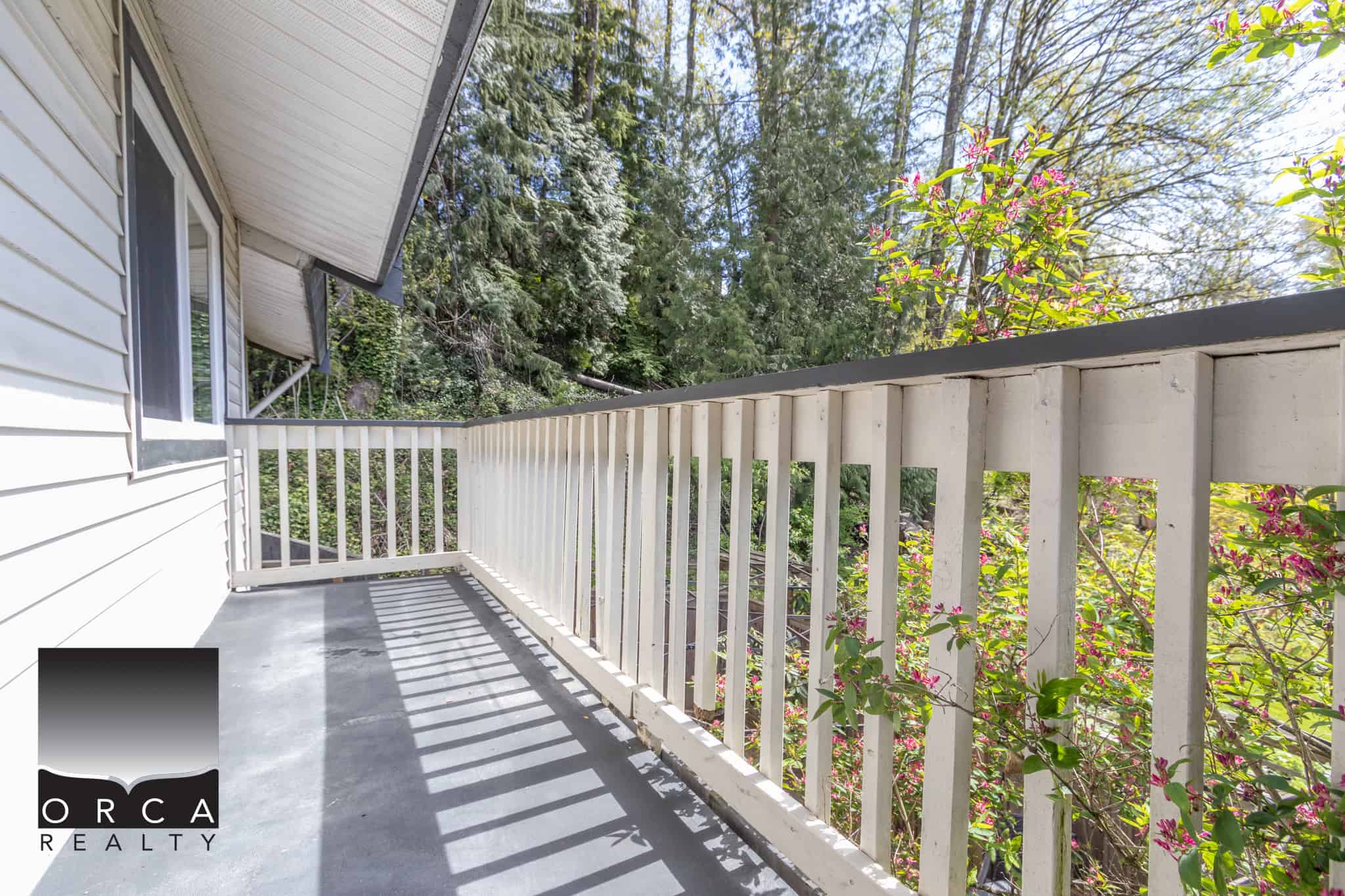 Balcony with white railing surrounded by lush greenery in a residential property listing by Orca Realty Inc.