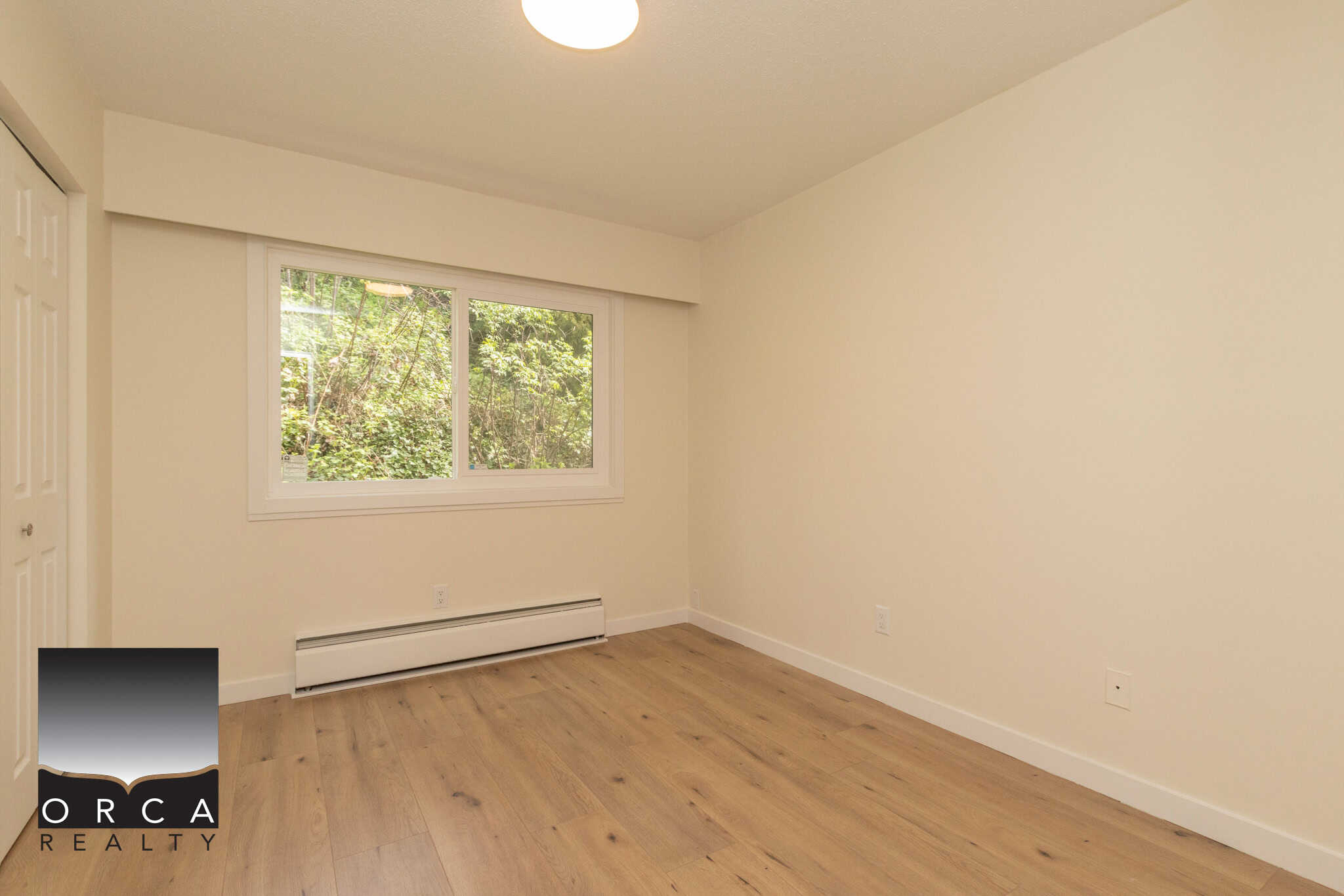 Bright empty bedroom with large window, neutral walls, hardwood flooring, and baseboard heating in a modern home for sale by Orca Realty Inc.