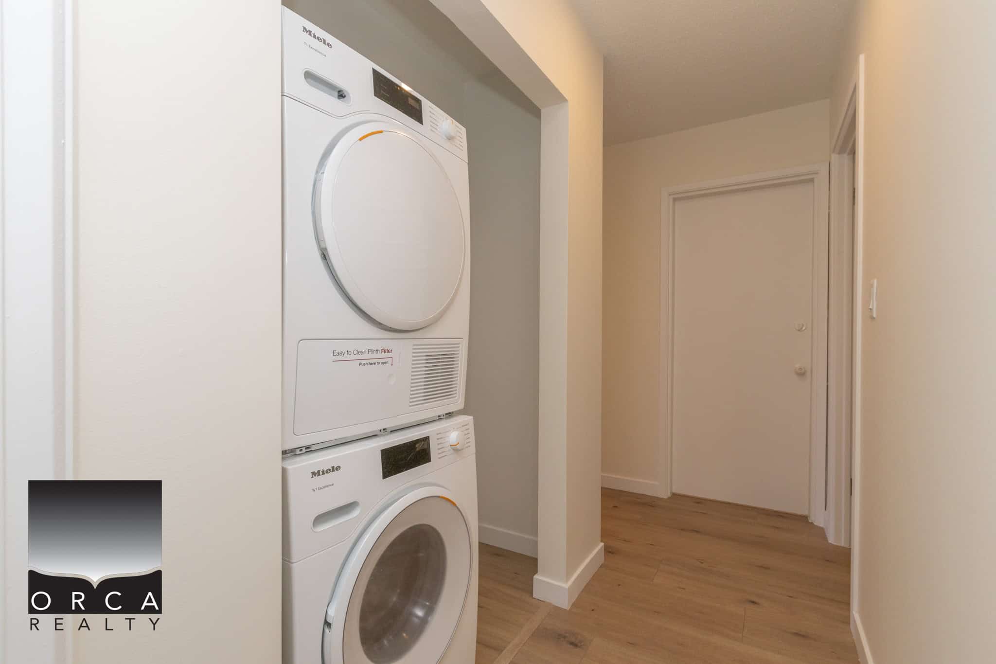 Stacked washer and dryer units in a laundry closet within a modern home, featuring white appliances and a wood floor setting.