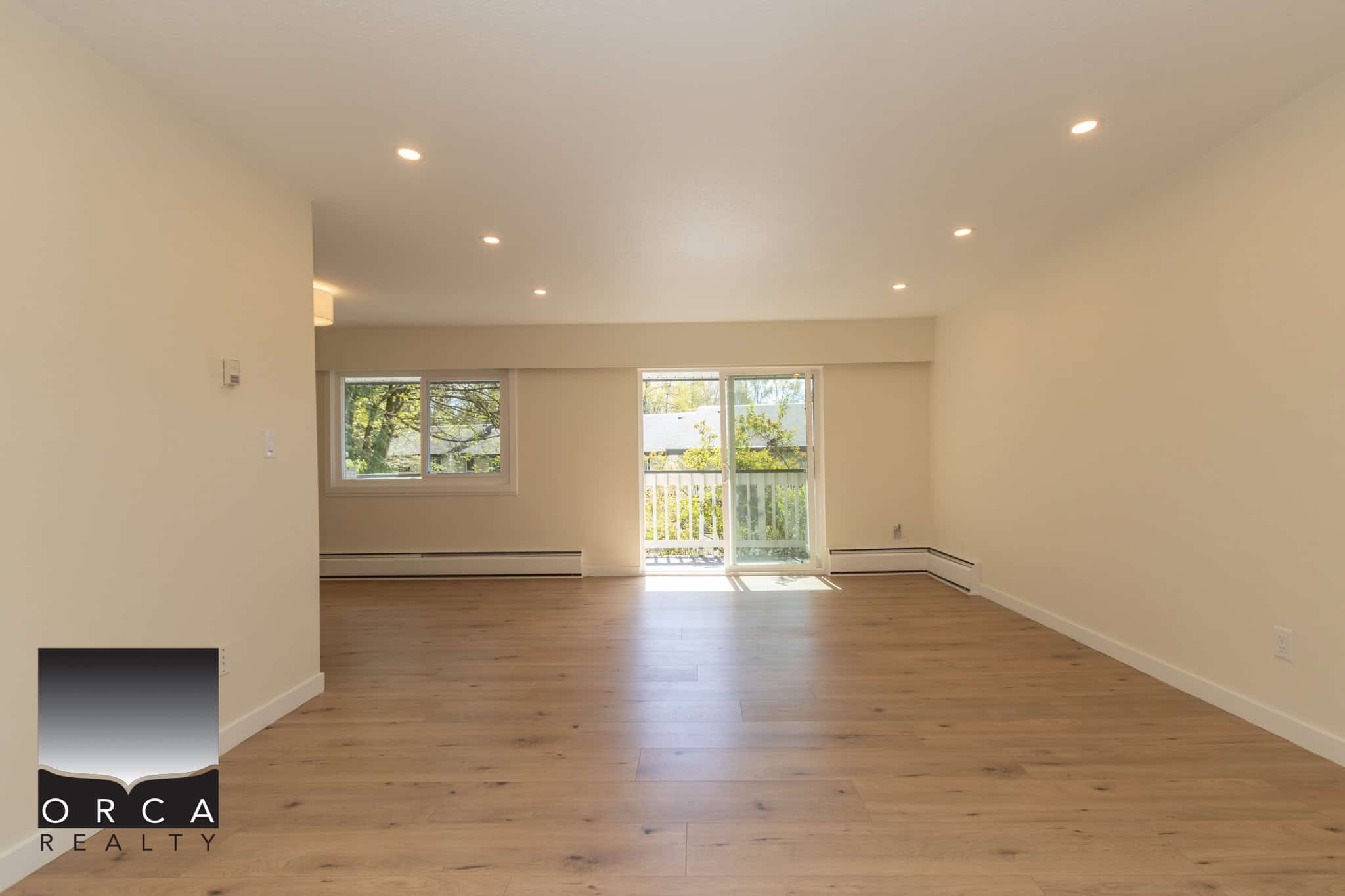 Bright and spacious living room with large windows, hardwood flooring, and access to balcony, ideal for Vancouver Island real estate buyers seeking modern efficiency and natural light.