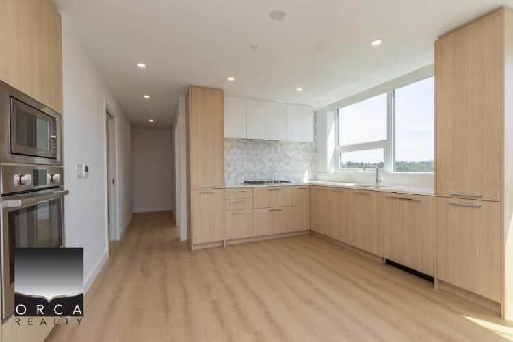 Modern kitchen with light wood cabinetry, large windows, and stainless steel appliances in a new condo or apartment in Vancouver, BC.