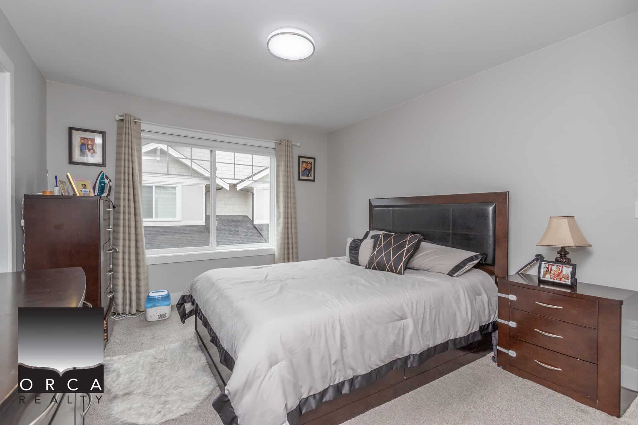 17 Modern bedroom featuring a large window, wooden bed frame with black leather headboard, cozy bedding, bedside table with lamp, and neutral decor – ideal for homebuyers in the Orca Realty real estate listings.