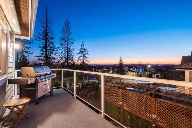 Spacious balcony with stunning sunset views over a suburban neighbourhood, perfect for relaxing or entertaining. Includes a barbecue grill, wooden chair, and glass railing showcasing scenic property surroundings.