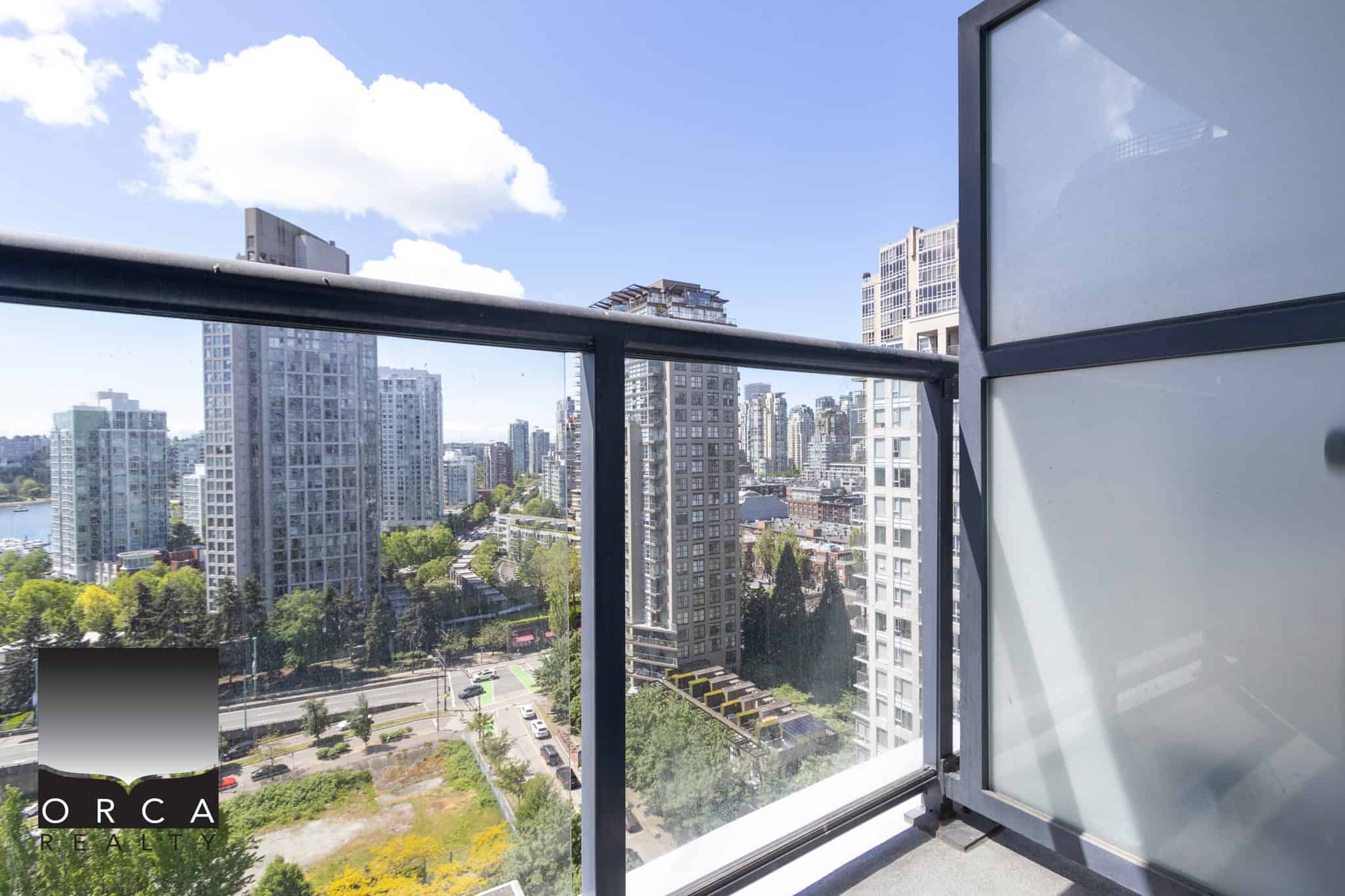 Modern city skyline view from a high-rise apartment balcony in Vancouver, featuring glass railing and urban landscape with tall buildings, lush greenery, and a bright blue sky.