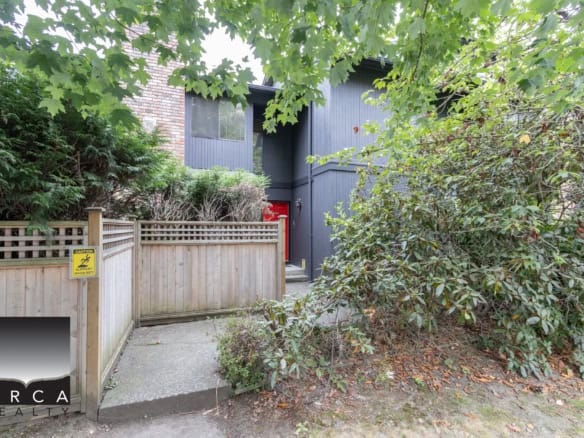 A modern townhouse exterior with a dark blue facade, surrounded by lush green trees and bushes, featuring a small concrete pathway and a wooden fence in a residential area in Vancouver, BC.