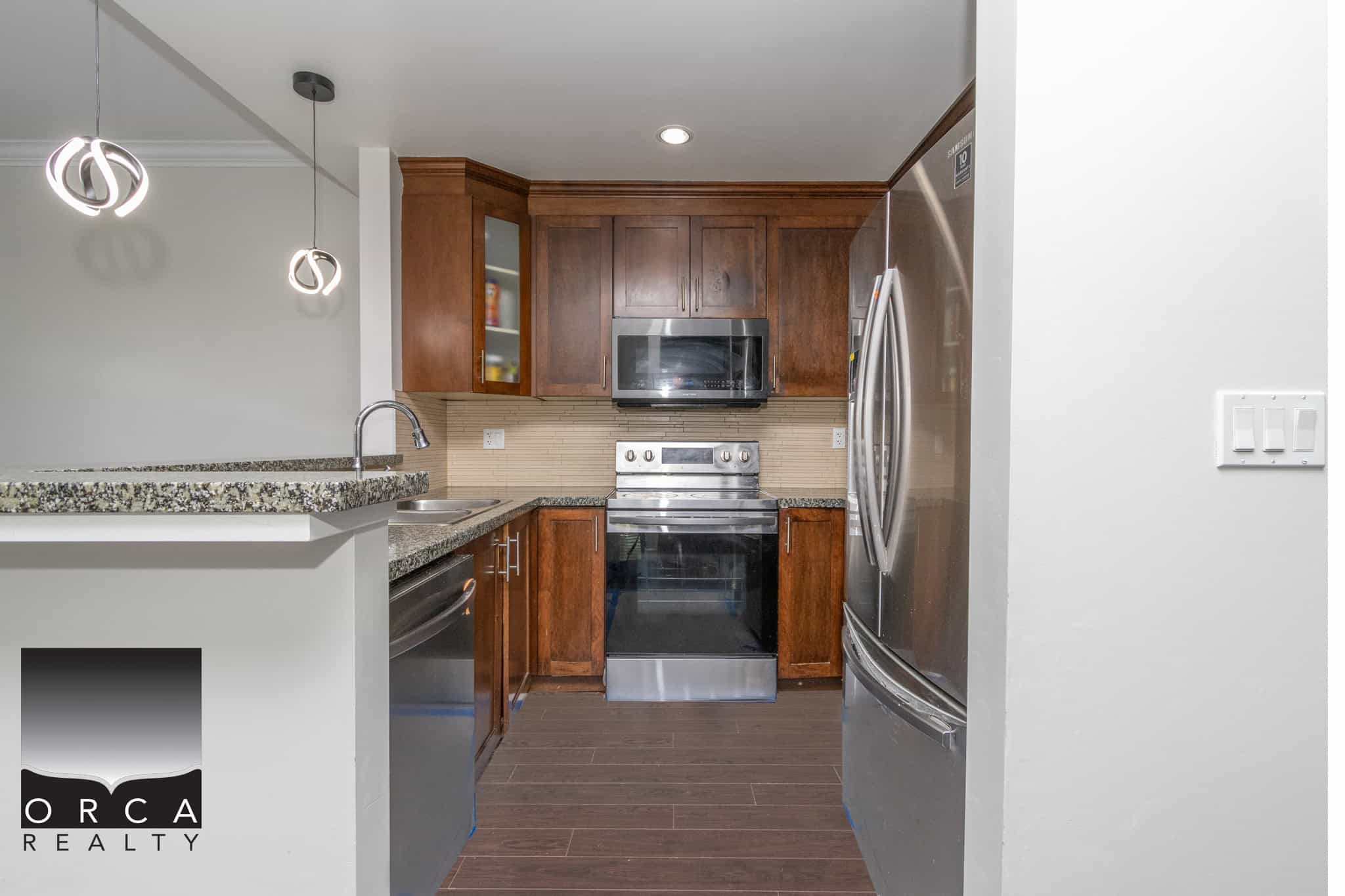 Stainless steel refrigerator and oven in modern kitchen with dark wood cabinetry and granite countertops, part of Orca Realty Inc.'s property listings in British Columbia.