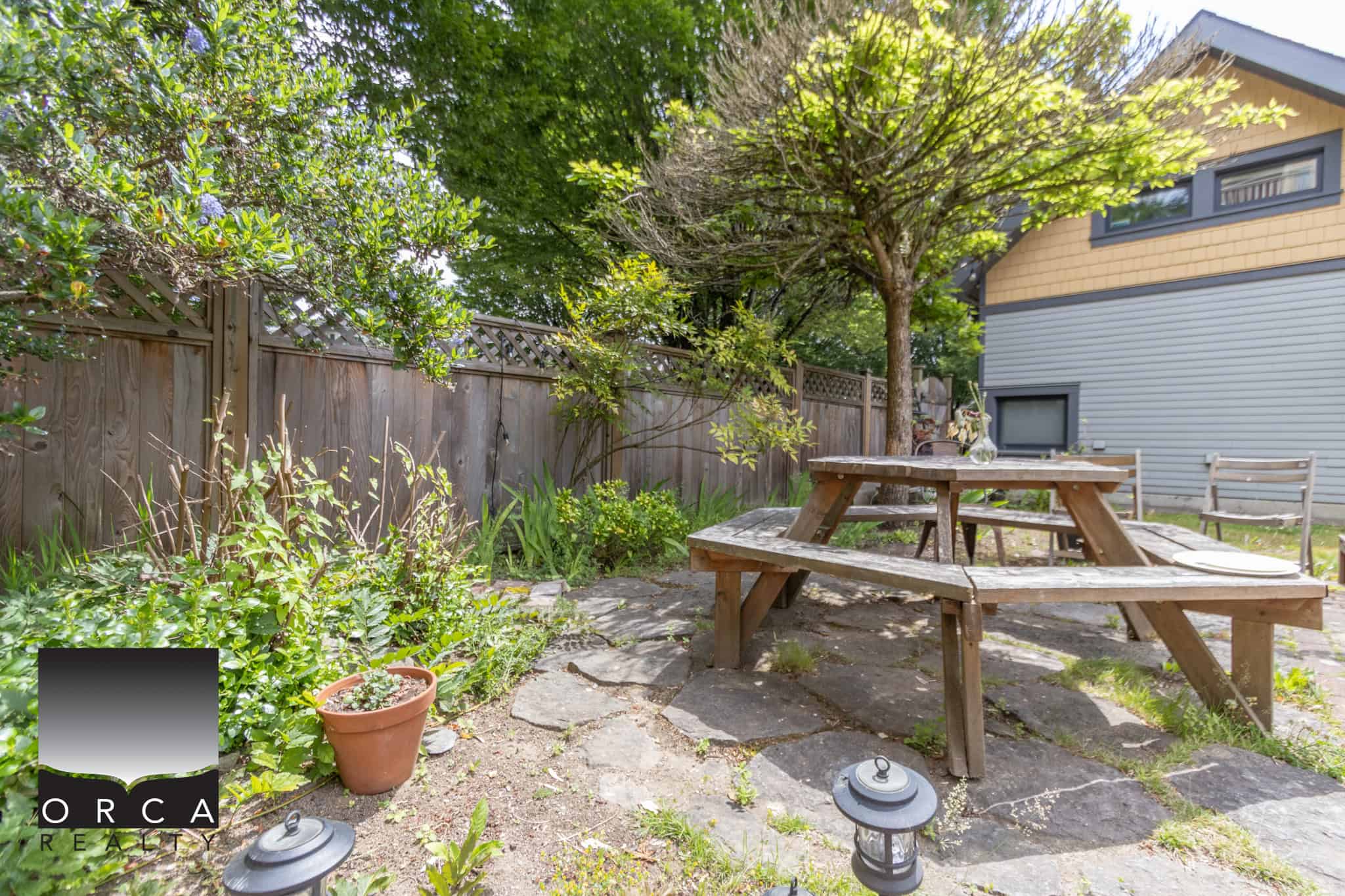 Charming backyard patio with wooden picnic table, surrounded by lush greenery, mature trees, and a weathered wooden fence, perfect for outdoor dining and relaxation.