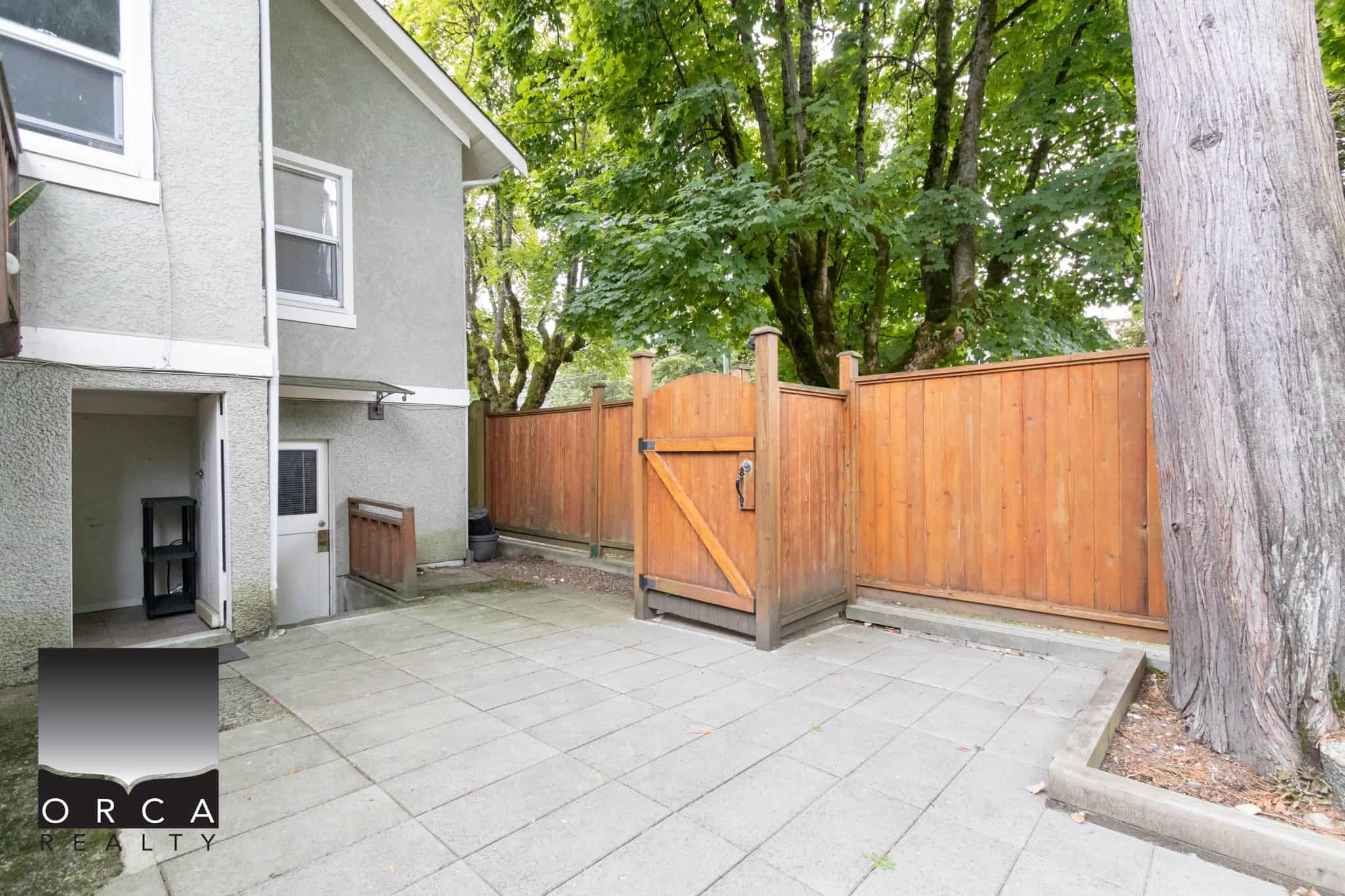Bright outdoor patio space with a wooden fence, mature trees, and a grey multi-family building, perfect for outdoor entertaining and enjoying nature in Victoria BC.