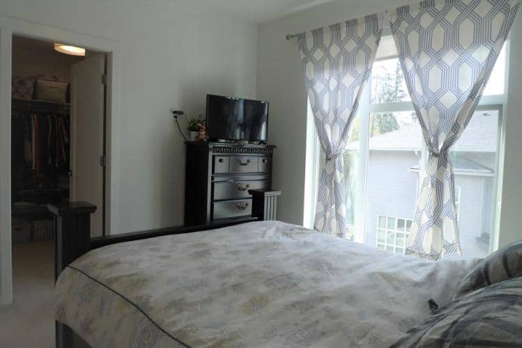 Modern bedroom with large windows and natural light, featuring a comfy bed and a black dresser with a flat-screen TV, perfect for a relaxed coastal home.
