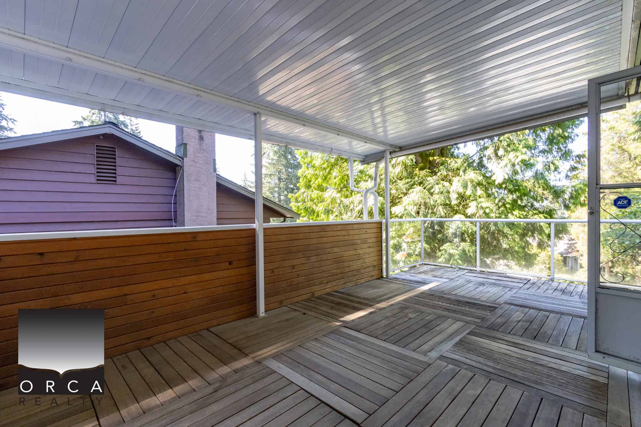 Spacious outdoor balcony with wooden flooring and a protective overhang, ideal for relaxing or entertaining, surrounded by lush green trees in a residential neighbourhood.