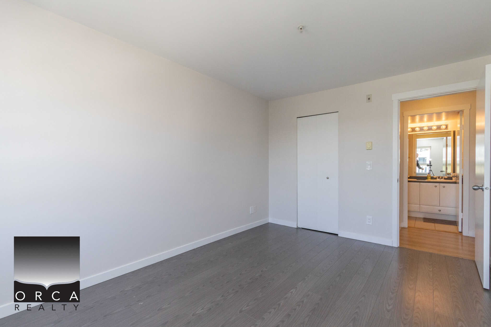 Bright empty modern bedroom with grey hardwood floors, white walls, and an open door leading to a bathroom, showcasing a clean, contemporary real estate interior property in Vancouver.