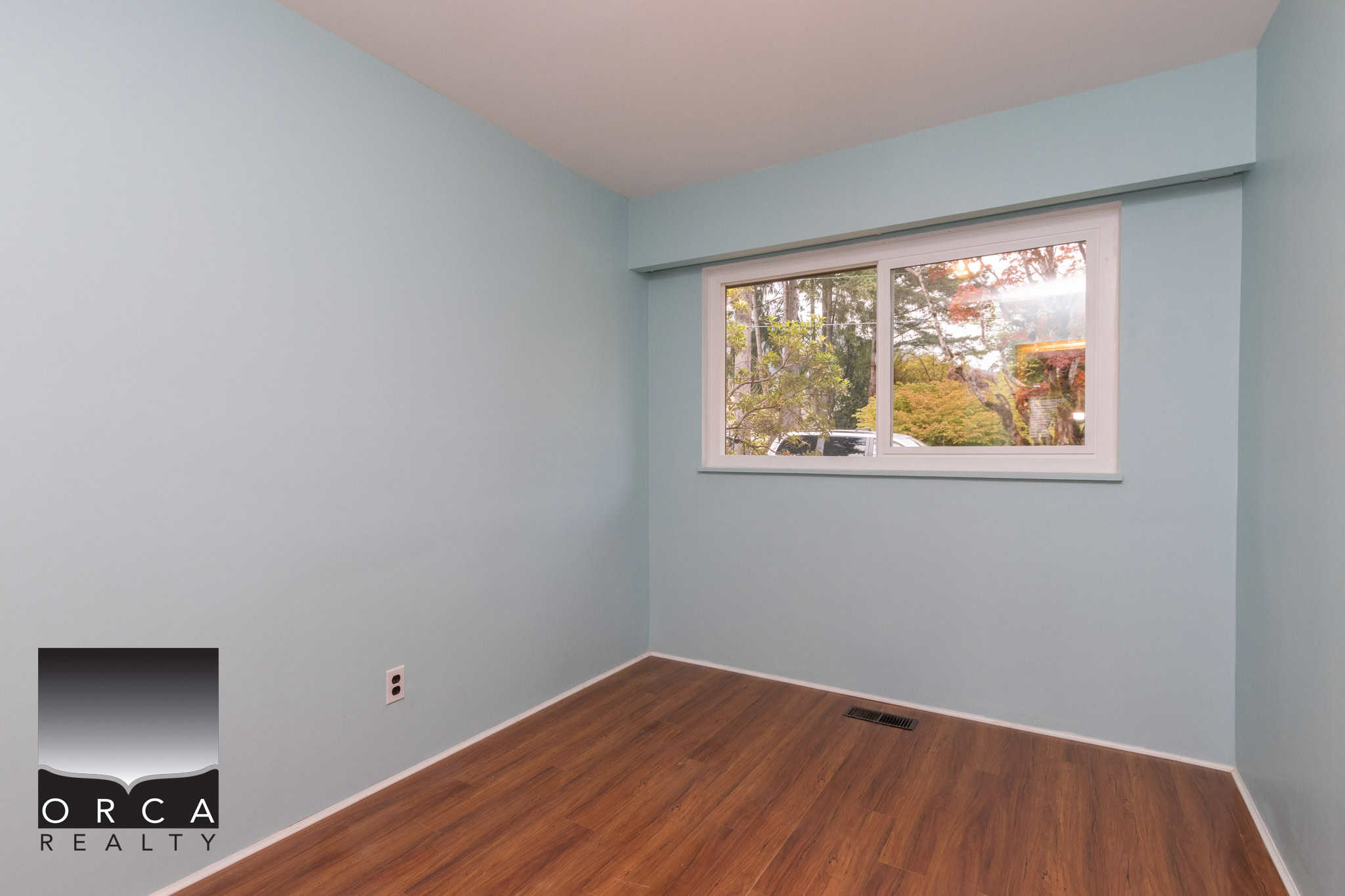 13 Bright, empty bedroom with light blue walls and natural wooden flooring, showcasing a large window with scenic greenery view, ideal for homebuyers seeking a comfortable and stylish space.