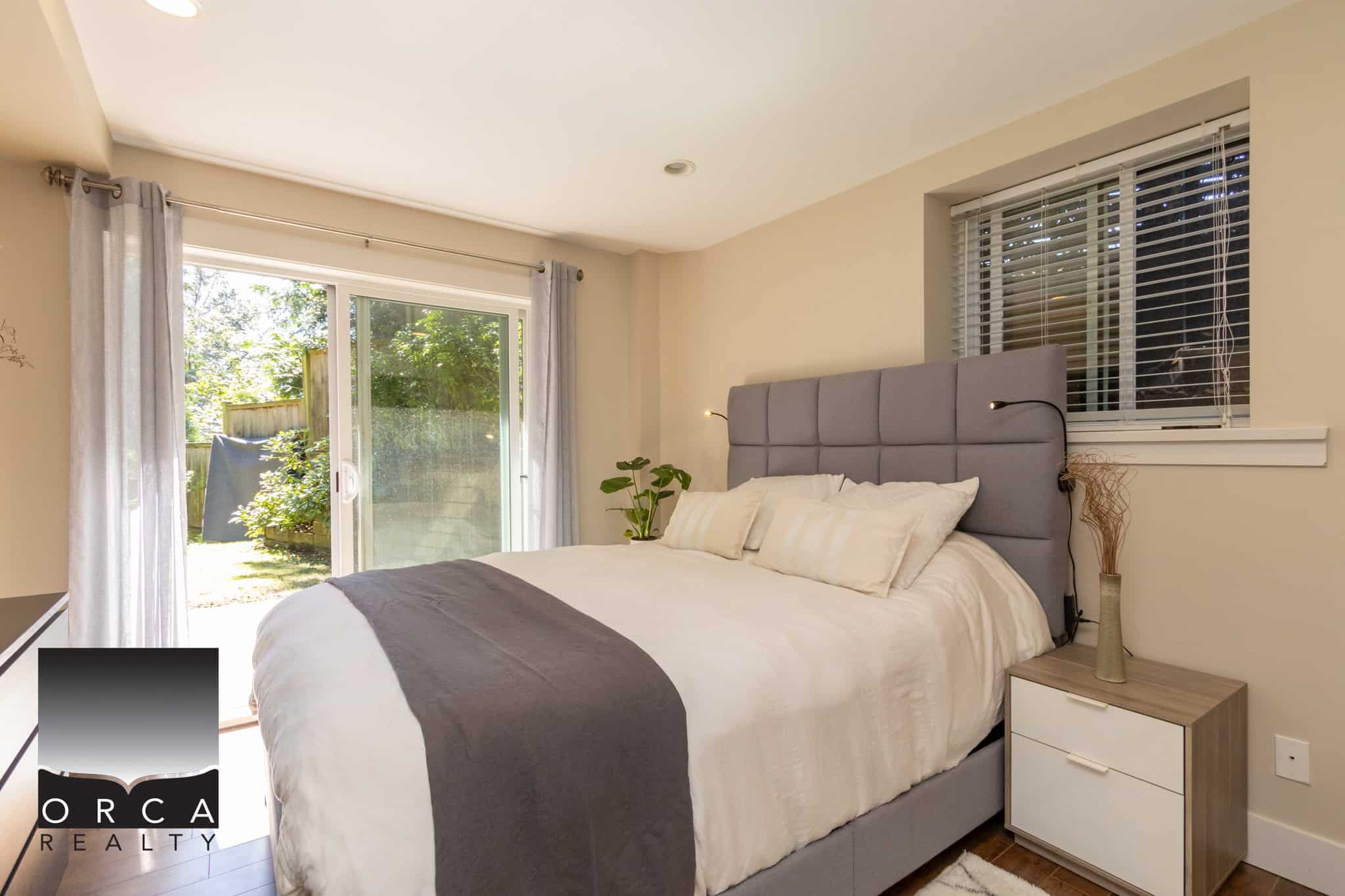 Cozy bedroom with a modern grey upholstered headboard, white bedding, and natural light from sliding glass door and window, ideal for your Vancouver Island home.