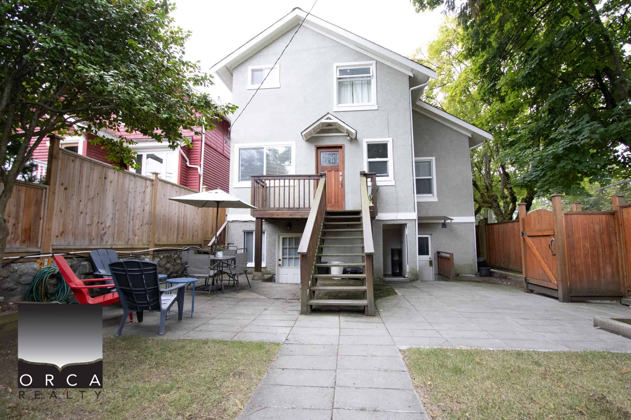 Bright backyard patio with outdoor seating and umbrella, featuring a multi-story grey house with wooden stairs, surrounded by mature trees and privacy fencing, ideal for family gatherings and outdoor relaxation.