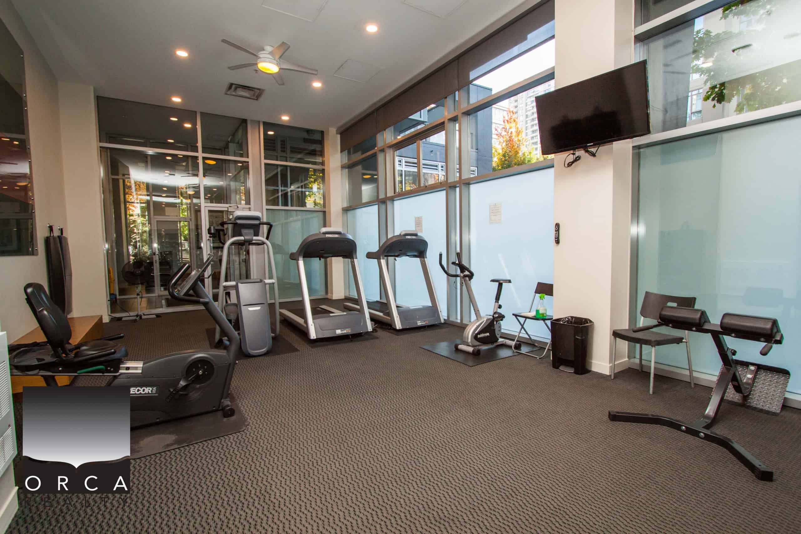 Modern urban gym with cardio equipment, large windows, and natural light, perfect for residents to maintain fitness routines within Orca Realty Inc. community premises.