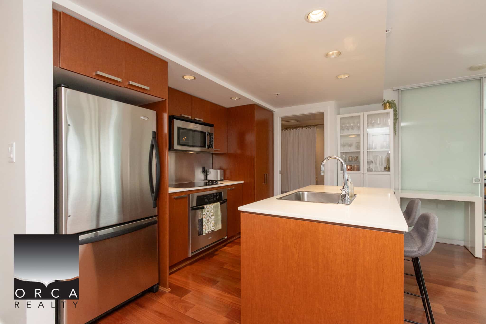 Stainless steel refrigerator and modern kitchen with warm wood cabinetry and a spacious island, ideal for cooking and entertaining in a stylish Orca Realty Inc. property in Vancouver.