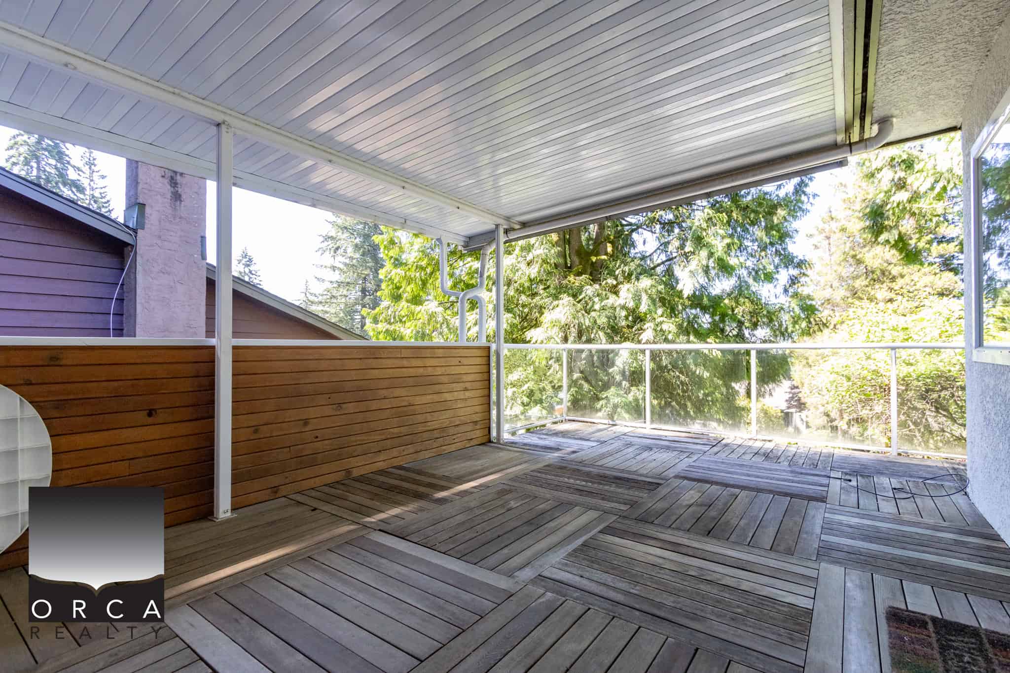 Balcony with wooden decking, glass railing, and lush green trees in the background, showcasing outdoor space for relaxation in Vancouver homes listed by Orca Realty Inc.