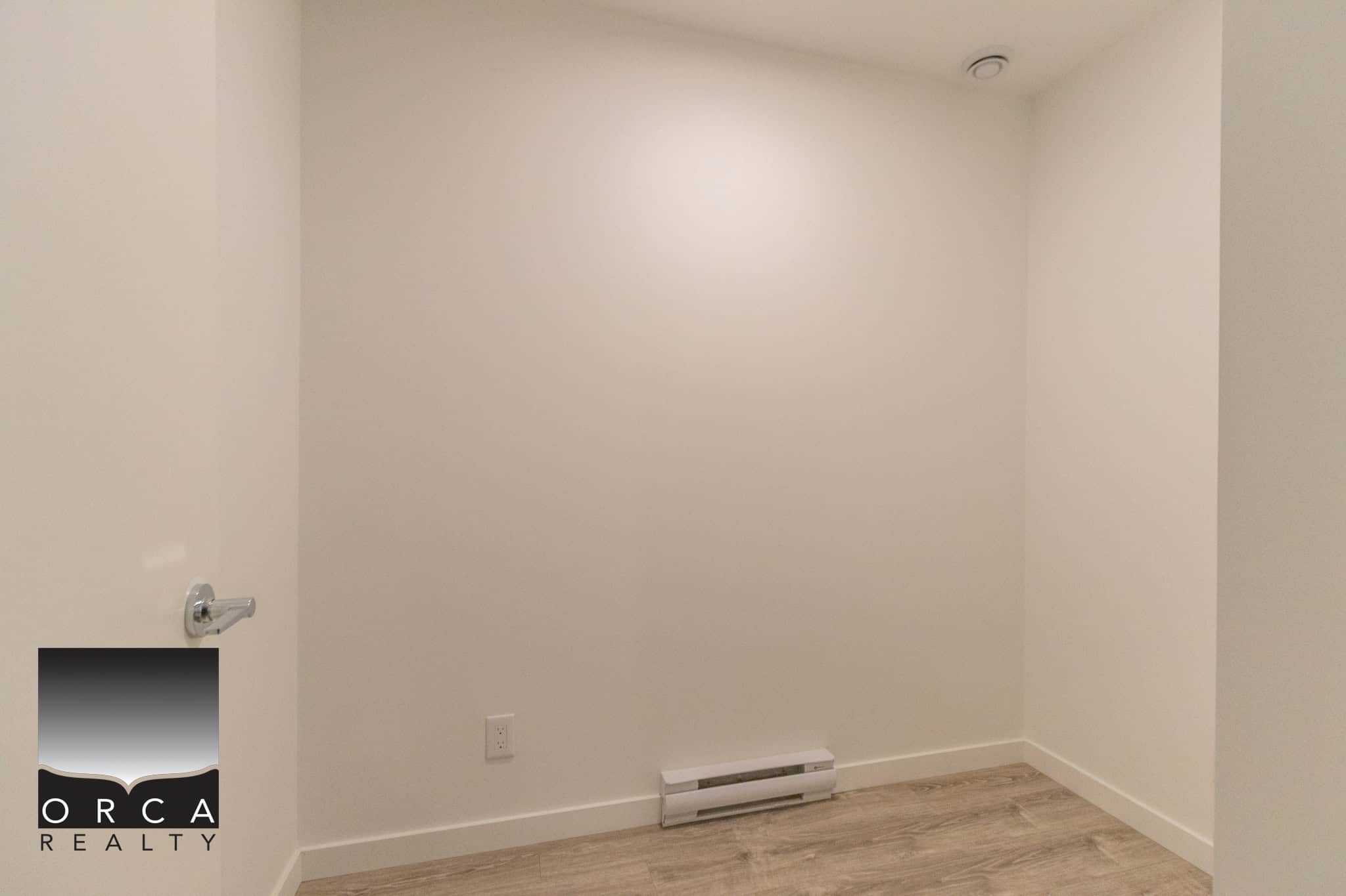 Bright empty room with white walls, wooden flooring, and baseboard heater, ideal for a modern home or rental property in Victoria, BC. Perfect for real estate listings to showcase a versatile interior space.