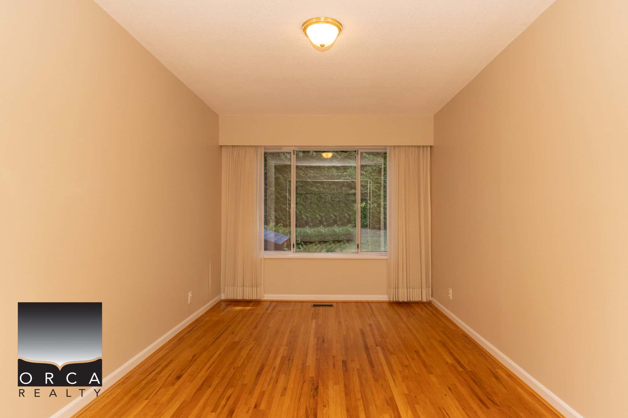 Bright empty living room with hardwood floors and a large window with beige curtains, ideal for home staging and real estate listings.