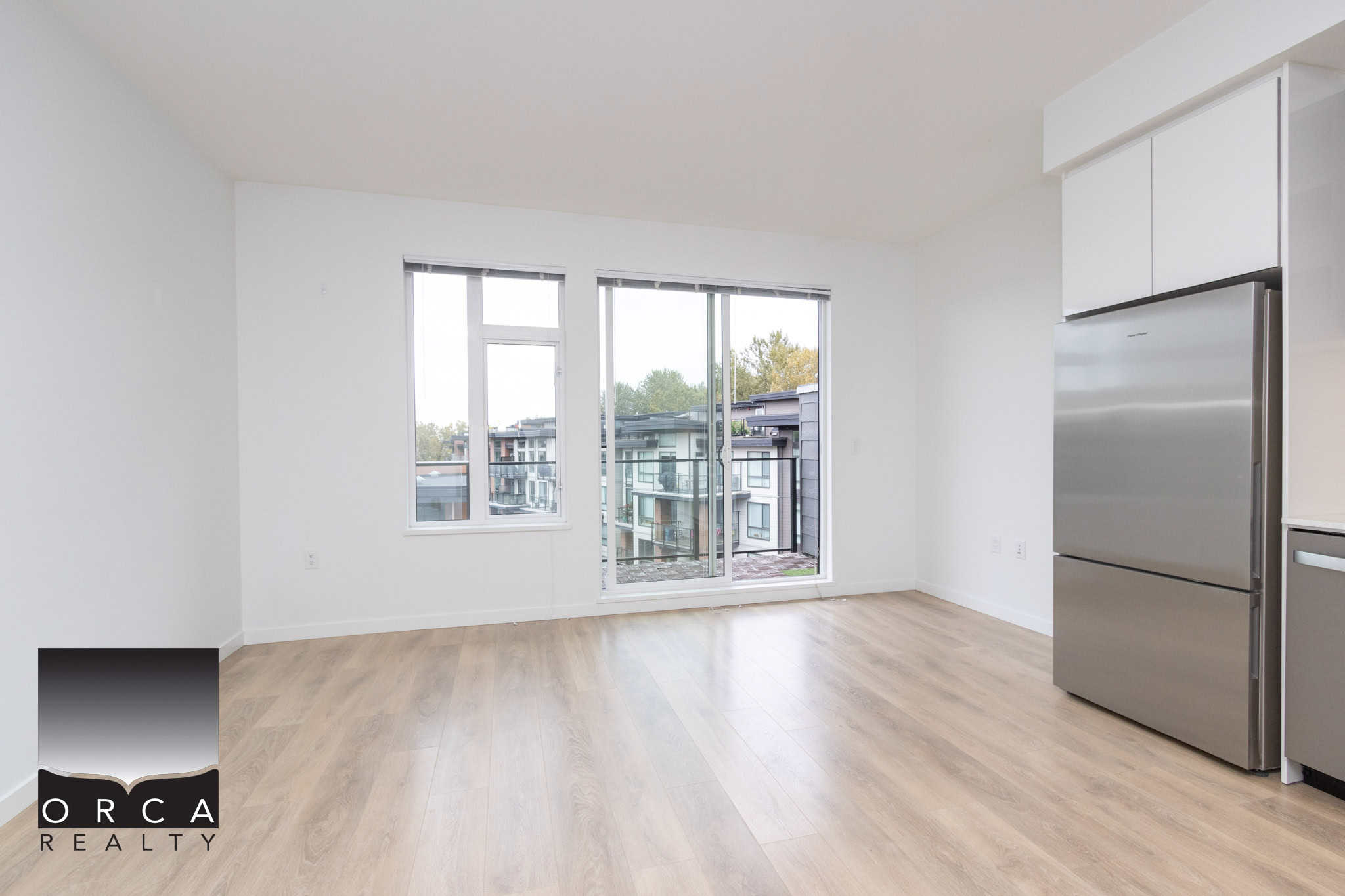Bright modern apartment living space with large windows, white walls, and wood flooring, featuring a stainless steel refrigerator, ideal for Vancouver area real estate.