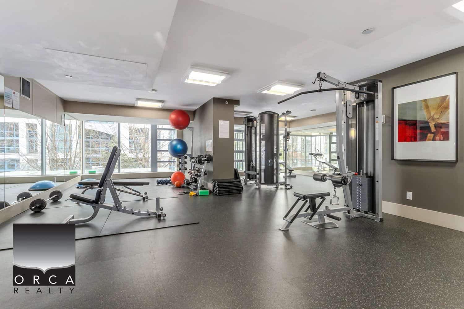 Modern fitness center with cardio machines, free weights, stability balls, and exercise equipment. Bright, spacious workout area with large windows and contemporary decor for residents’ health and wellness.