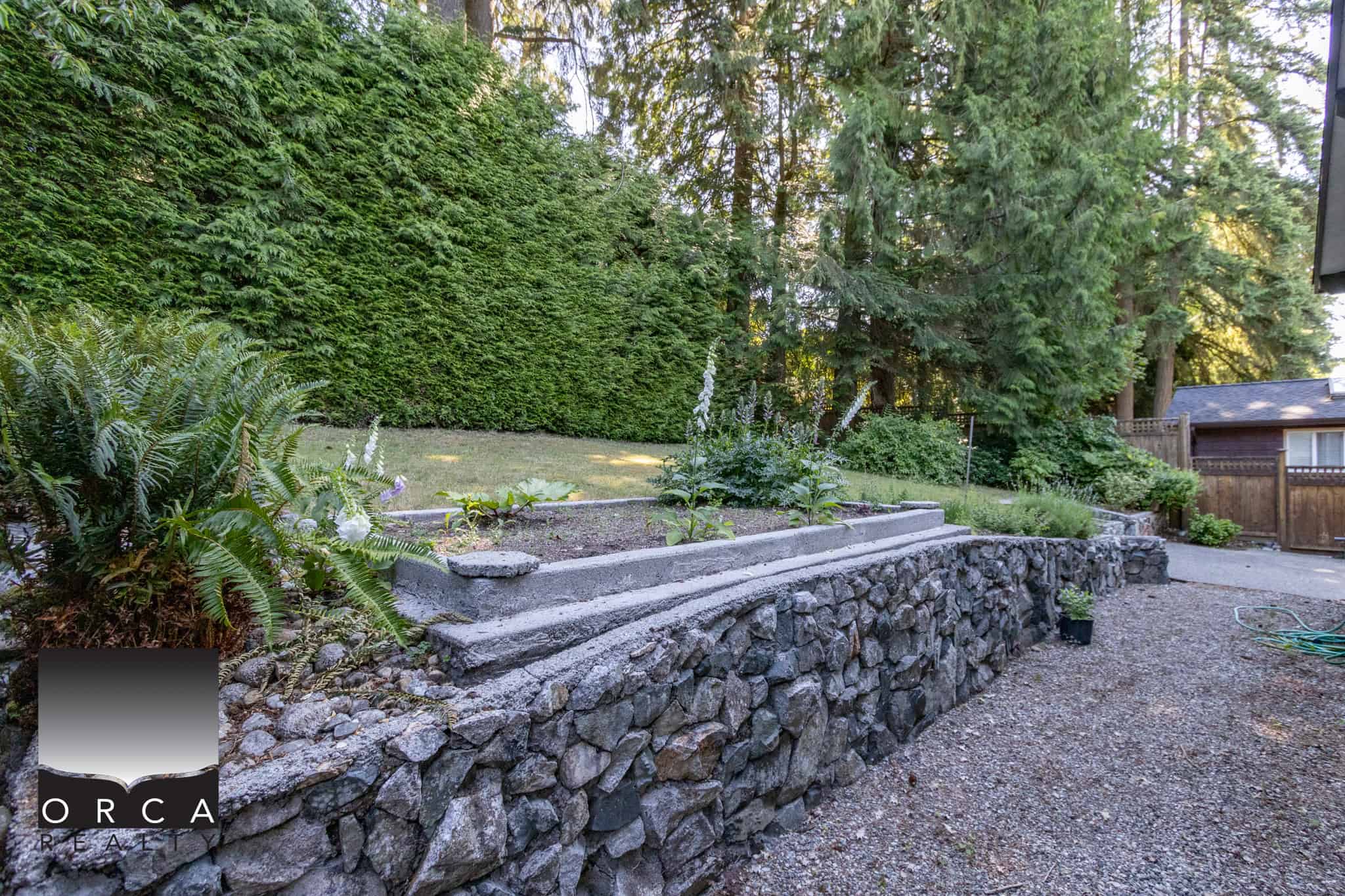 Lush backyard with stone retaining wall, garden beds, mature trees, and private outdoor space – perfect for Vancouver Island real estate buyers.