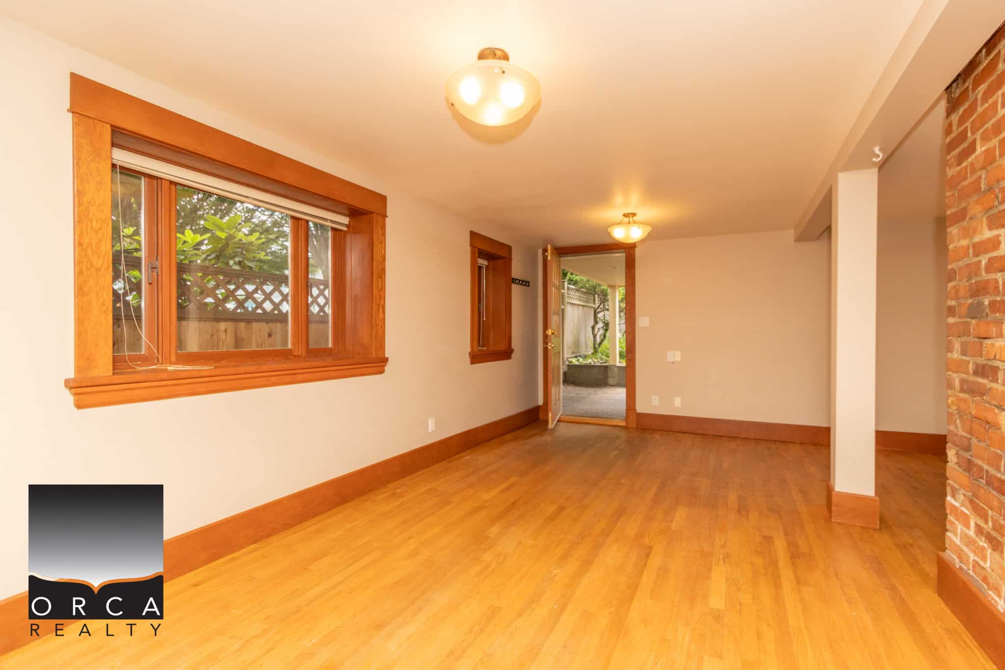 Bright living room with hardwood floors and large windows, featuring natural light, wooden trim, and brick accent wall, ideal for Vancouver Island real estate buyers.