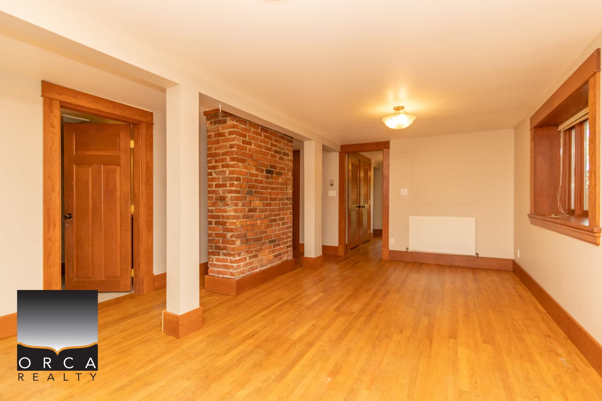Bright spacious living room with hardwood floors, exposed brick accent wall, and large windows offering natural light in this Orca Realty property in Victoria, BC.