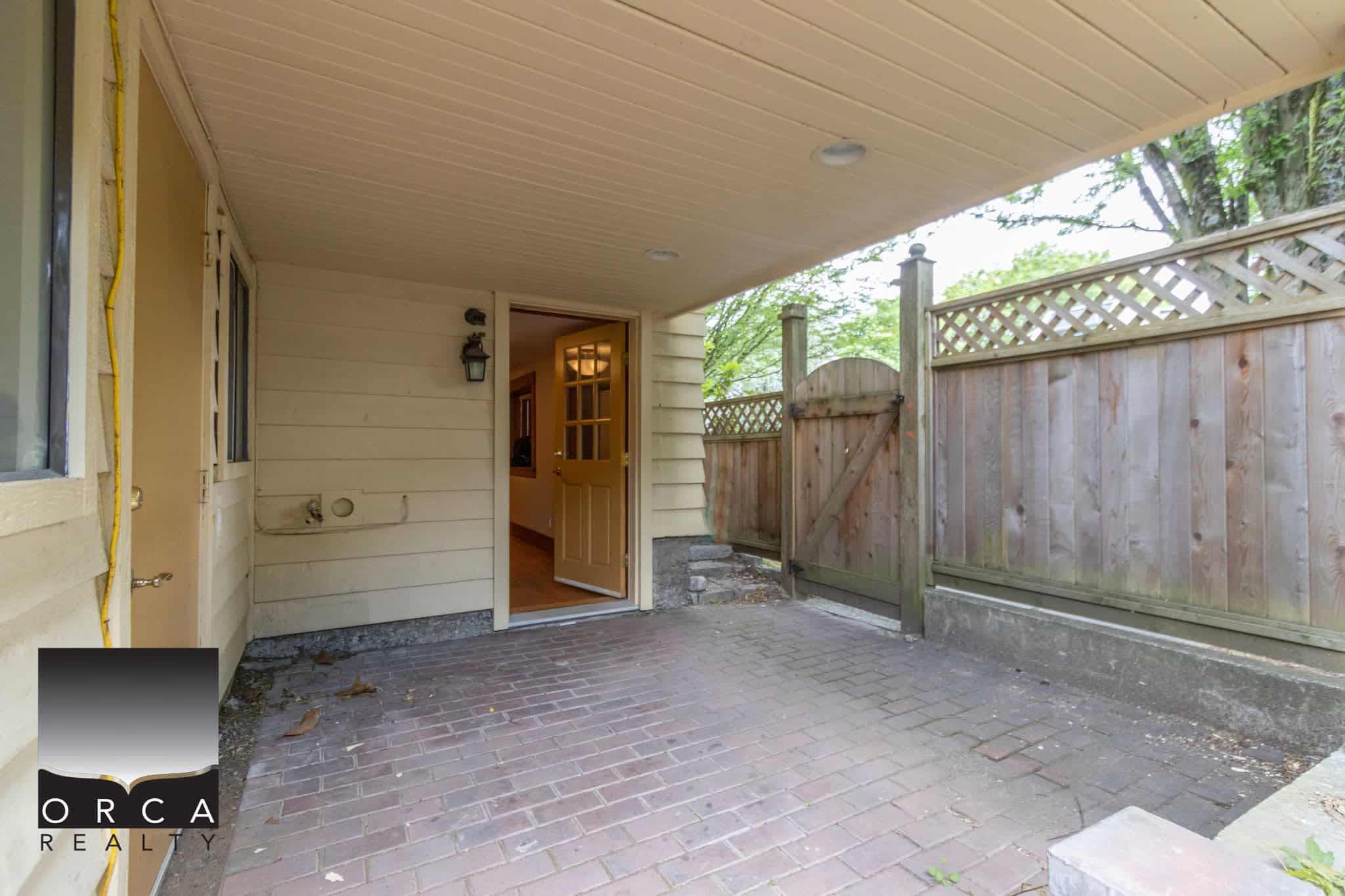 Bright covered patio area with brick flooring, leading to a home entrance in a peaceful neighbourhood, perfect for outdoor lounging, surrounded by wooden fencing for privacy.