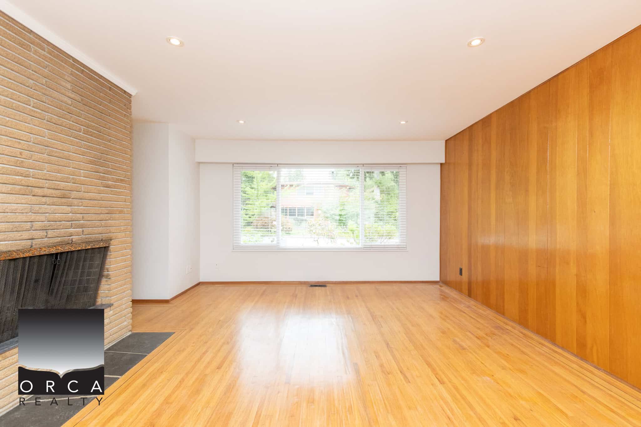 20 Spacious living room with natural light, hardwood floors, brick fireplace, and large window, showcasing Orca Realty Inc.'s expertise in Vancouver Island real estate.