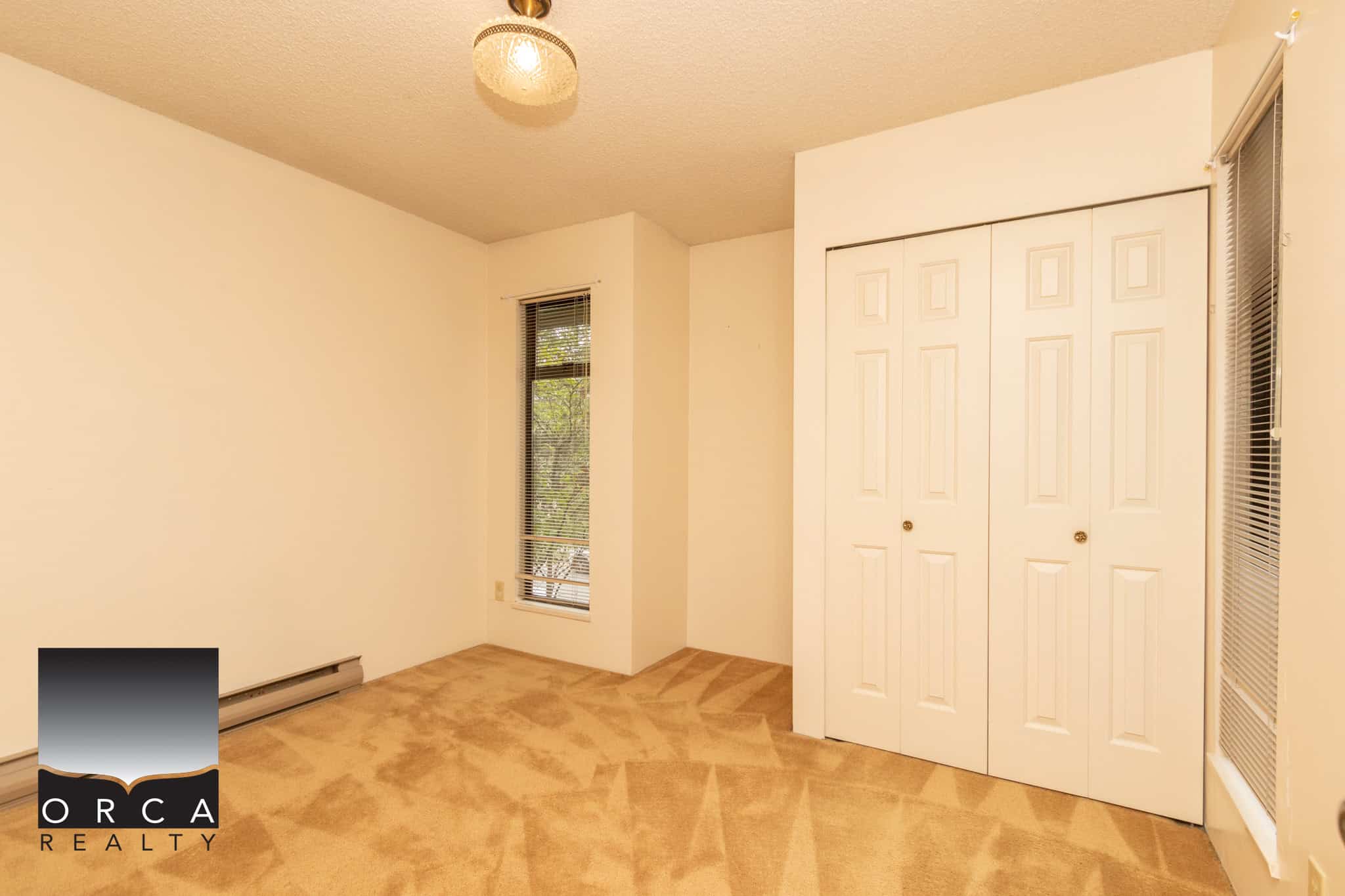 Bright empty bedroom with beige carpet, white walls, large window with blinds, and double closet doors, showcasing a clean and inviting space for residential real estate in Vancouver.