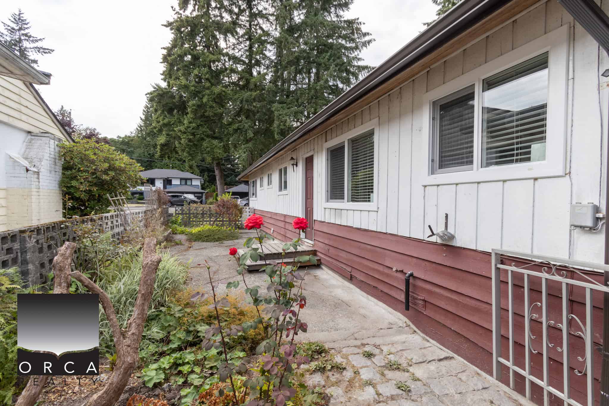 21 Cozy single-story home exterior with a garden, white siding, window blinds, and a paved walkway, available through Orca Realty Inc., ideal for buyers seeking a Vancouver Island property.