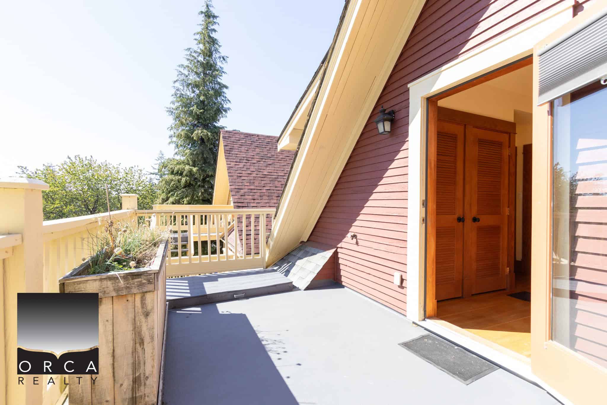 Spacious outdoor balcony with wooden railing and planters, attached to a colourful red house with a sliding glass door, perfect for relaxing and enjoying views in Vancouver's real estate market.