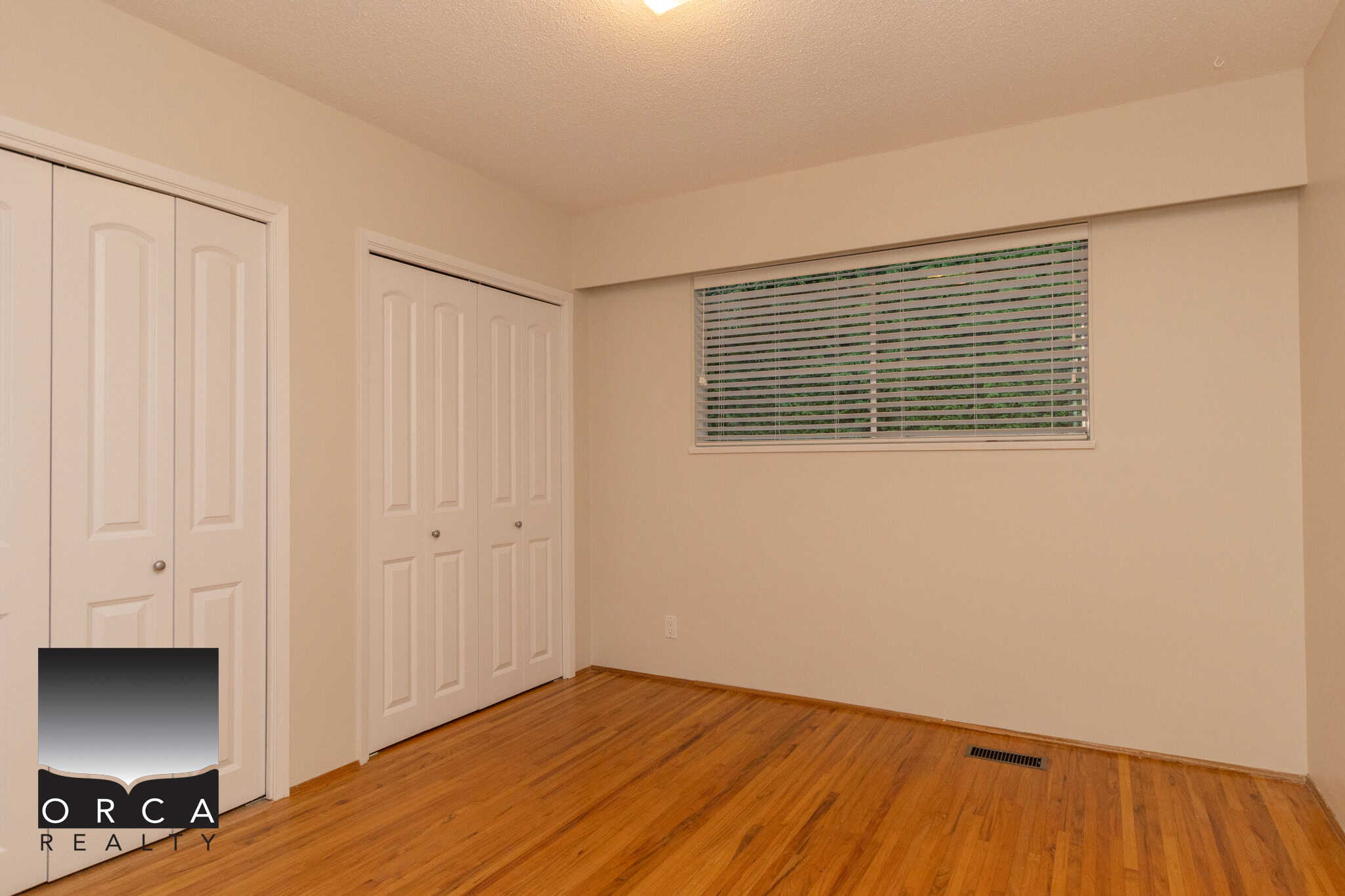 Bright empty bedroom with white closet doors, hardwood flooring, and large window with blinds in a Toronto house listed by Orca Realty Inc.